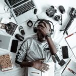 A young man lying on a bed surrounded by modern gadgets, books, and tech devices, illustrating digital lifestyle.